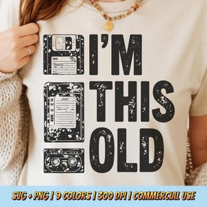 May include: Beige t-shirt with a distressed black graphic that reads "I'M THIS OLD." The design includes illustrations of a floppy disk, cassette tape, and a compact cassette. The shirt is a retro design.