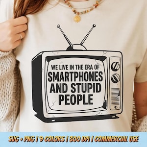Peut inclure: T-shirt beige avec un dessin noir et blanc d'un ancien téléviseur. Le texte sur l'écran dit "WE LIVE IN THE ERA OF SMARTPHONES AND STUPID PEOPLE." Le t-shirt a un col rond et des manches courtes.