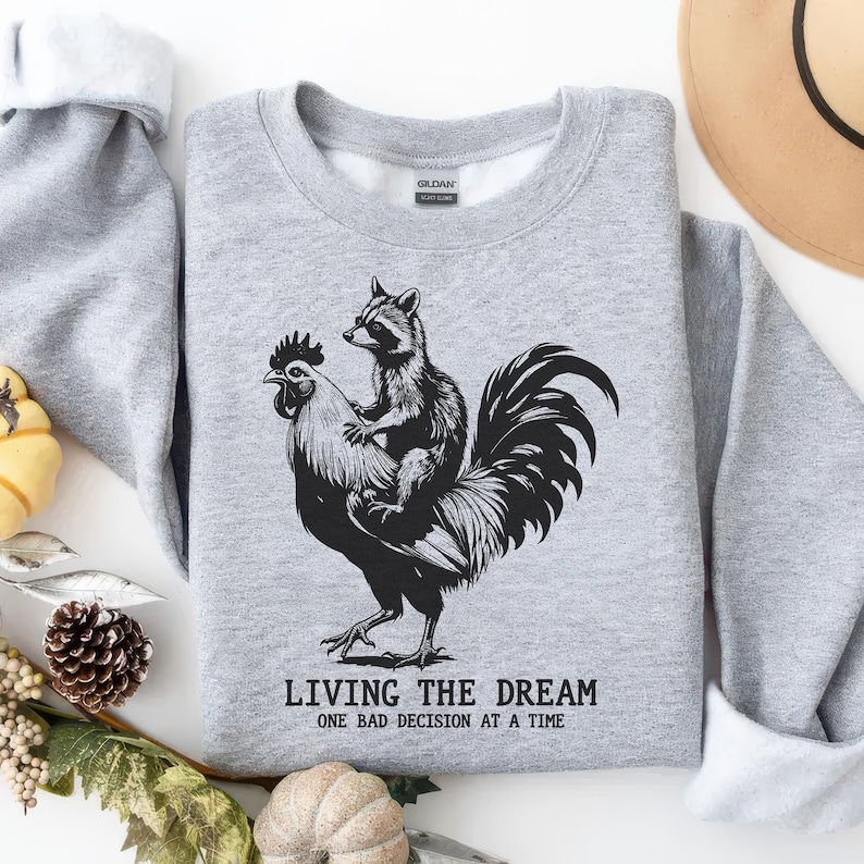 May include: A heather gray sweatshirt featuring a black and white graphic of a raccoon riding a rooster. The text "LIVING THE DREAM ONE BAD DECISION AT A TIME" is printed below the graphic. The sweatshirt has a crew neck and long sleeves.