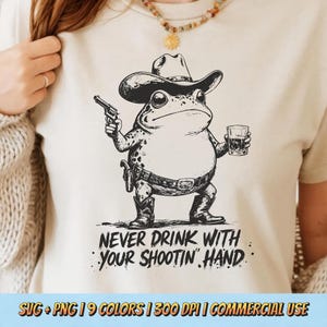 Puede incluir: Camiseta color crema con una rana de dibujos animados con sombrero de vaquero, sosteniendo una pistola y un vaso de whisky. El texto "NEVER DRINK WITH YOUR SHOOTIN' HAND" está impreso debajo. El diseño es en blanco y negro.