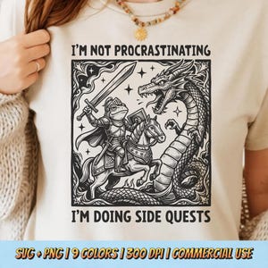 May include: Cream-colored t-shirt featuring a black and white illustration of a frog knight on horseback, facing a dragon. Text reads "I'M NOT PROCRASTINATING I'M DOING SIDE QUESTS". The design includes stars and decorative borders.