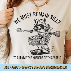 Pode incluir: Camiseta bege com um desenho preto de um guaxinim com armadura de cavaleiro em um carrinho de compras, segurando um tridente e uma vassoura. O texto diz "WE MUST REMAIN SILLY" e "TO SURVIVE THE HORRORS OF THIS WORLD."