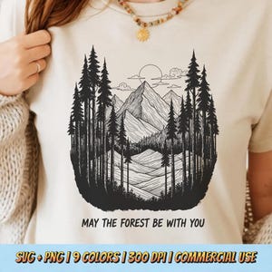 以下が含まれることがあります： ベージュのTシャツ。黒と白の森林と山のグラフィックが特徴です。デザインには、高い木、山、太陽、そして「MAY THE FOREST BE WITH YOU」というテキストが含まれています。