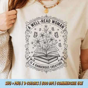Puede incluir: Una camiseta beige con un gráfico en blanco y negro. El gráfico incluye un libro abierto con flores y mariposas, y el texto "A WELL-READ WOMAN IS A DANGEROUS CREATURE". La camiseta es de color neutro.