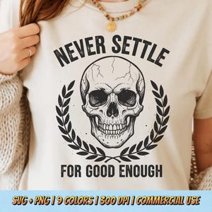 Può includere: Una maglietta color crema con una dettagliata illustrazione di un teschio circondato da una corona d'alloro. Il testo "NEVER SETTLE" è sopra il teschio, con "FOR GOOD ENOUGH" sotto. Il design è nero.