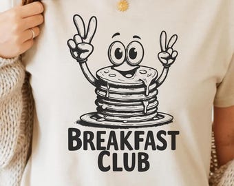 Könnte beinhalten: Ein cremefarbenes T-Shirt zeigt einen schwarz-weißen Cartoon-Pfannkuchenstapel mit einem lächelnden Gesicht und Friedenszeichen. Der Text "BREAKFAST CLUB" befindet sich unter der Grafik. Der untere Teil des Shirts hat den Text "SVG + PNG | 9 COLORS | 300 DPI | COMMERCIAL USE".