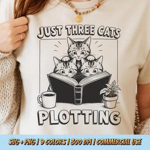 May include: A cream-colored t-shirt featuring a black graphic of three cats reading an open book. The text above reads JUST THREE CATS and below reads PLOTTING. A coffee mug and a small potted plant are positioned on either side of the book.