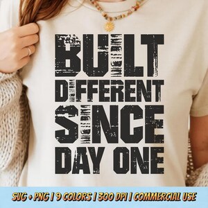Op de afbeelding: Een beige t-shirt met de tekst "BUILT DIFFERENT SINCE DAY ONE" in verweerde zwarte letters. De onderkant van het shirt toont ook de tekst "SVG + PNG | 9 COLORS | 300 DPI | COMMERCIAL USE".