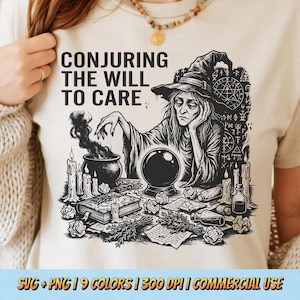May include: Beige t-shirt featuring a black and white illustration of a witch with a crystal ball, cauldron, and candles. The text "CONJURING THE WILL TO CARE" is above the illustration. The design has a mystical theme.