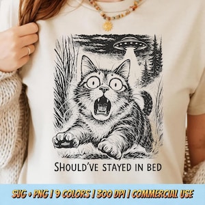 May include: Beige t-shirt featuring a black and white illustration of a startled cat being beamed up by a UFO. The text "SHOULD'VE STAYED IN BED" is below the graphic. The shirt is a light neutral color.