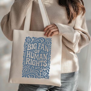Puede incluir: Bolso tote de color crema con el texto "BIG FAN OF HUMAN RIGHTS" en azul, rodeado de dise&ntilde;os florales y de volutas. El bolso tiene correas largas de color crema.