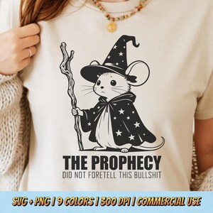 May include: A cream-colored t-shirt featuring a black and white illustration of a mouse dressed as a wizard, holding a staff. The text "THE PROPHECY DID NOT FORETELL THIS BULLSHIT" is printed below the illustration.