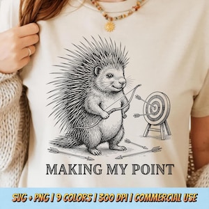 May include: Beige t-shirt featuring a detailed black and white illustration of a porcupine holding a bow and arrow, aiming at a target. The text "MAKING MY POINT" is below the image. Arrows are scattered around the target.