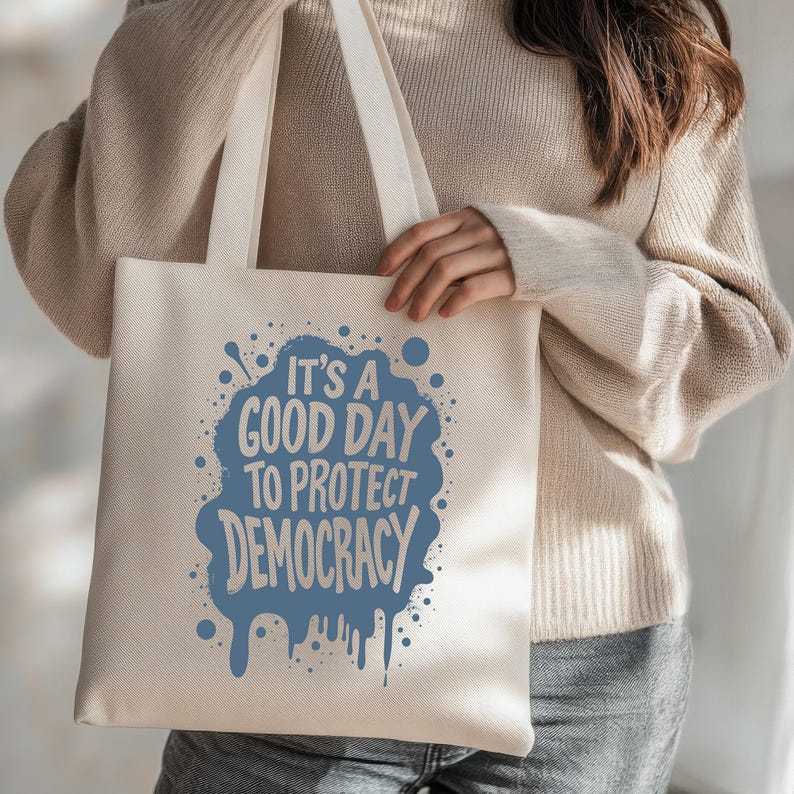 May include: Cream-colored tote bag with the text "It's a good day to protect democracy" in blue. The text is in a stylized font with a splatter design. The bag has long, cream-colored handles.