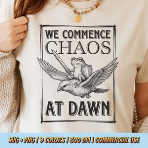 Könnte beinhalten: Beigefarbenes T-Shirt mit schwarzer Grafik. Die Grafik zeigt einen Frosch, der auf einem Vogel reitet und ein Schwert und einen Schild hält, mit dem Text "WE COMMENCE CHAOS AT DAWN". Das T-Shirt hat eine neutrale Farbe.