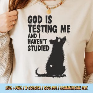 May include: Beige t-shirt with the text "GOD IS TESTING ME AND I HAVEN'T STUDIED" in black. A black rat illustration is below the text, looking upwards. The bottom of the image has text that reads "SVG + PNG | 9 COLORS | 300 DPI | COMMERCIAL USE."