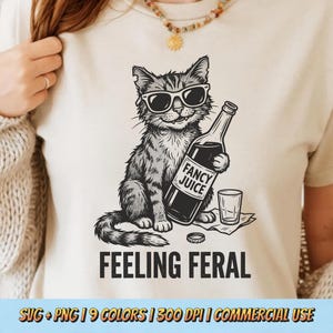 以下が含まれることがあります： ベージュのTシャツ。サングラスをかけた猫が「FANCY JUICE」と書かれたボトルを持っている白黒イラスト。猫の下には「FEELING FERAL」の文字。グラスとボトルキャップも描かれています。