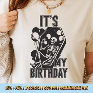 May include: Beige t-shirt featuring a black graphic of a skeleton reclining in a coffin, with balloons. The text reads "IT'S MY BIRTHDAY". The design is suitable for Halloween or birthday celebrations.
