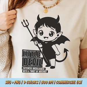 May include: A cream-colored t-shirt featuring a black and white cartoon devil with a trident, horns, and a tail. The text "LITTLE DEVIL" is above "CUTENESS OVERLOADED WITH A TWIST OF MISCHIEF". The shirt is worn by a person.