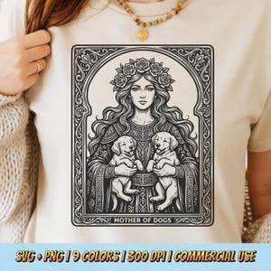 May include: Beige t-shirt featuring a black and white illustration of a woman with a floral crown holding two puppies. The image is framed with ornate details and the text "MOTHER OF DOGS" is at the bottom. The shirt has a crew neck.