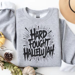 May include: A heather gray sweatshirt with the words "HARD FOUGHT HALLELUJAH" in bold, black lettering. The text is surrounded by a burst design. The sweatshirt is folded, with the sleeves and collar visible. A fall-themed arrangement is in the background.