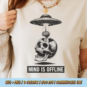 May include: Beige t-shirt featuring a black and white graphic of a UFO beaming a brain into a skull. The text "MIND IS OFFLINE" is below the graphic. The shirt has a crew neck and short sleeves.
