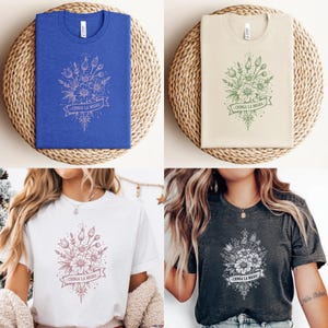 May include: Four t-shirts in different colors: blue, beige, white, and charcoal gray. Each shirt features a floral design with the text "CHINGA LA MIGRA" on a banner. The shirts are displayed on a woven, circular surface.
