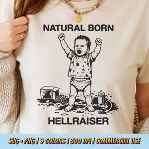 May include: Cream-colored t-shirt with the words "NATURAL BORN" above a graphic of a baby with arms raised and the word "HELLRAISER" below. The baby is wearing a diaper and a dotted shirt. A box, bottle, and cup are also in the graphic.