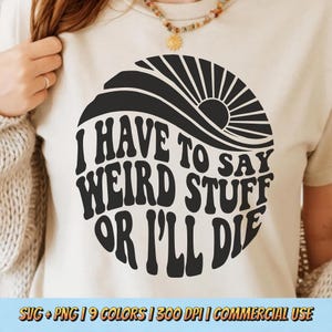 May include: A cream-colored t-shirt with a black circular graphic. The graphic features the text "I HAVE TO SAY WEIRD STUFF OR I'LL DIE" in a retro font, with a sun design. The shirt is a neutral color.