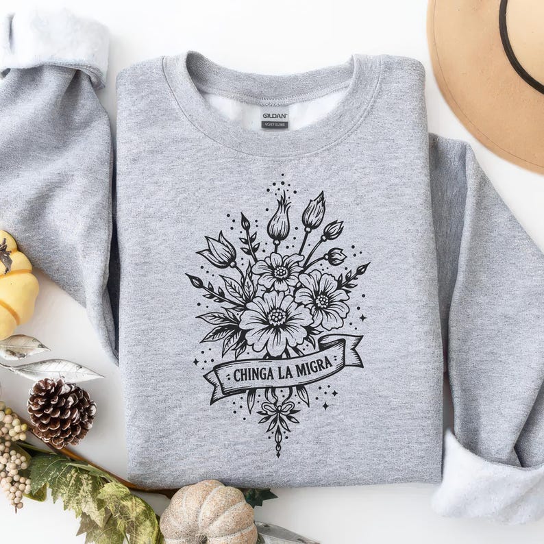 May include: A heather gray sweatshirt with a floral design in black ink. The design features a bouquet of flowers, buds, and leaves, with a banner that reads "CHINGA LA MIGRA". The sweatshirt has a crew neck and long sleeves.