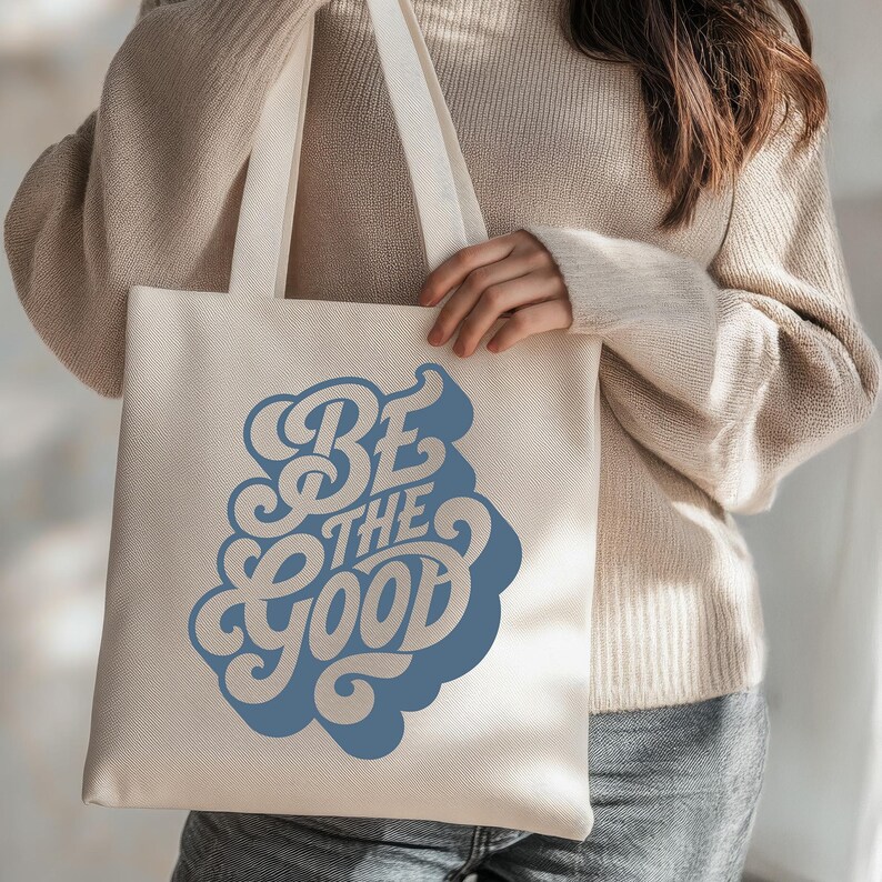 May include: Cream-colored tote bag with the phrase "BE THE GOOD" in a retro blue font. The bag has long straps and is made of a sturdy fabric, suitable for carrying groceries, books, or other items. The design is simple and eye-catching.