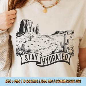 May include: Beige t-shirt featuring a black and white desert landscape design with cacti and mesas. The text "STAY HYDRATED" is displayed on a banner across the bottom. The shirt is a neutral color.