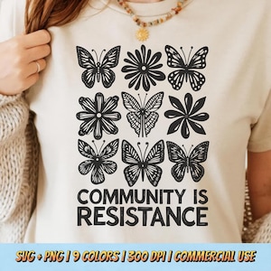 以下が含まれることがあります： クリーム色のTシャツ。黒いグラフィックデザインで、蝶と花が格子状に配置されています。「COMMUNITY IS RESISTANCE」というフレーズが、デザインの下に太字の黒い文字で印刷されています。柔らかい素材で作られたシャツです。