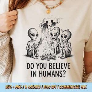 Puede incluir: Camiseta beige con un diseño en blanco y negro de tres extraterrestres alrededor de una fogata. El texto "DO YOU BELIEVE IN HUMANS?" está debajo. Un extraterrestre sostiene una linterna.