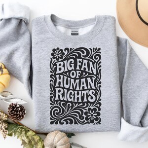 Puede incluir: Una sudadera gris claro con el texto negro "BIG FAN OF HUMAN RIGHTS" en una fuente decorativa, rodeada de elementos florales y arremolinados. La sudadera est&aacute; doblada, revelando un cuello interior blanco. El dise&ntilde;o est&aacute; centrado.