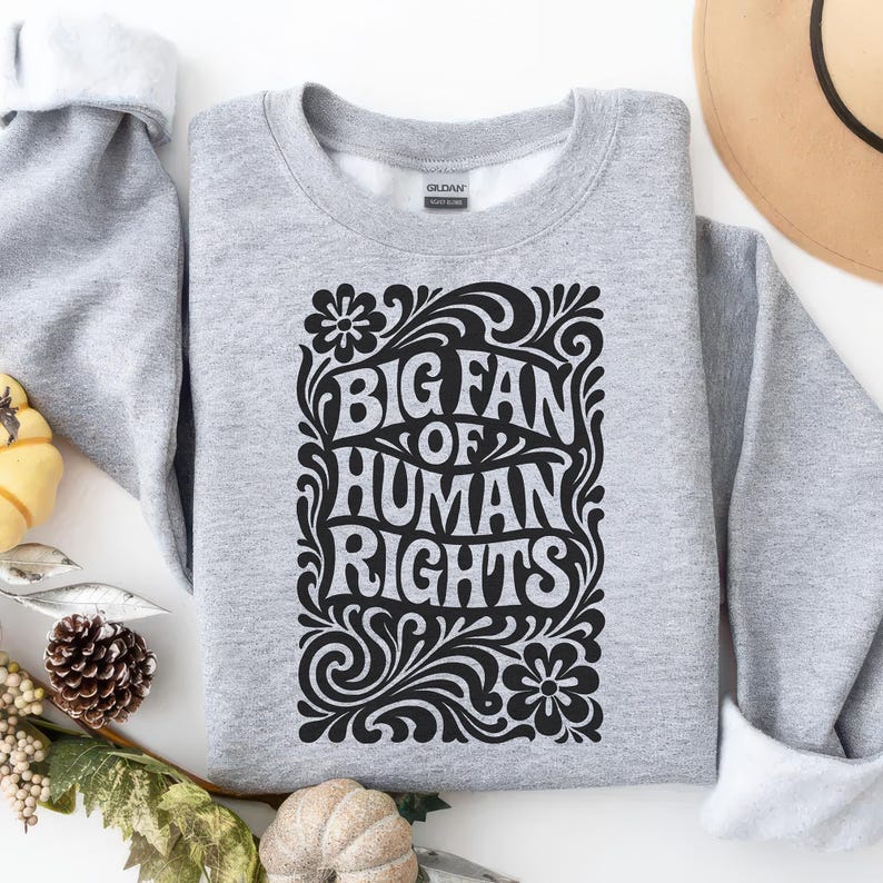 Puede incluir: Una sudadera gris claro con el texto negro "BIG FAN OF HUMAN RIGHTS" en una fuente decorativa, rodeada de elementos florales y arremolinados. La sudadera est&aacute; doblada, revelando un cuello interior blanco. El dise&ntilde;o est&aacute; centrado.