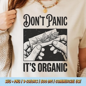 May include: Cream-coloured t-shirt featuring a black graphic with the text "DON'T PANIC IT'S ORGANIC". The graphic depicts hands rolling a cannabis joint. The shirt has a round neck and short sleeves.