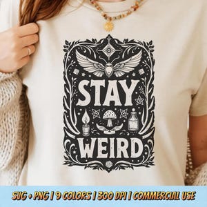 Puede incluir: Camiseta color crema con un diseño gráfico negro. El diseño presenta las palabras "STAY WEIRD" dentro de un marco decorativo, junto con un insecto alado, estrellas, una seta, una vela y una botella. La parte inferior de la camiseta tiene el texto "SVG + PNG | 9 COLORS | 300 DPI | COMMERCIAL USE".
