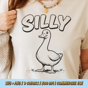 May include: Cream-colored t-shirt featuring a black and white illustration of a duck with the word "SILLY" above it in a playful font. The duck is smiling and standing on grass.