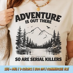 May include: Beige t-shirt with a black graphic of a mountain landscape and the text "ADVENTURE IS OUT THERE, BUT THEN AGAIN SO ARE SERIAL KILLERS." The design includes trees, water, and birds. The shirt is a neutral color.