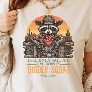 Pode incluir: Uma camiseta branca com um desenho animado de um guaxinim usando um chapéu de cowboy e uma bandana. O guaxinim está em pé na frente de uma cerca de madeira com cactos e um pôr do sol ao fundo. O texto na camiseta diz "The only squat I'm doin is diddly squat".
