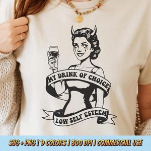 May include: A cream-colored t-shirt featuring a black and white illustration of a woman with devil horns holding a wine glass. The text "MY DRINK OF CHOICE" and "LOW SELF ESTEEM" are displayed on the shirt. The shirt has a round neckline.