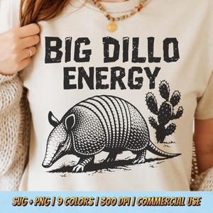 May include: A beige t-shirt with the text "BIG DILLO ENERGY" above a black and white illustration of an armadillo and a cactus. The shirt has a crew neck and short sleeves.