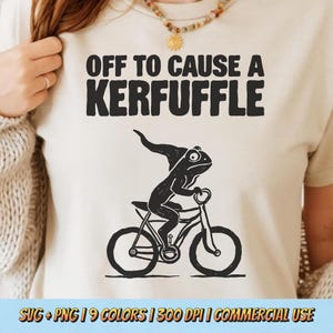 Puede incluir: Camiseta color crema con la frase negra "OFF TO CAUSE A KERFUFFLE". Debajo, una ilustración negra de una rana con sombrero montada en bicicleta. Prenda de vestir informal.