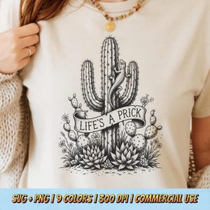 Peut inclure: T-shirt beige avec un motif de cactus noir et blanc, un lézard et le texte "LIFE'S A PRICK" sur une bannière. Le motif comprend des cactus, des plantes succulentes et des fleurs. Le t-shirt est de couleur neutre.