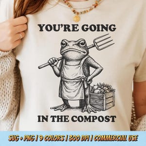 May include: Cream-coloured t-shirt featuring a black and white illustration of a frog in an apron, holding a pitchfork. The text above reads "YOU'RE GOING" and below reads "IN THE COMPOST".