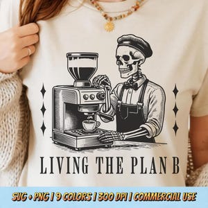 Op de afbeelding: Beige t-shirt met een zwart-witte illustratie van een skeletbarista die een espressomachine bedient. De tekst "LIVING THE PLAN B" staat eronder. Het ontwerp bevat ruitvormige decoraties.
