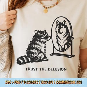 May include: A cream-colored t-shirt featuring a black and white illustration of a raccoon looking into a mirror, seeing a wolf. The text "TRUST THE DELUSION" is below the image. Additional text at the bottom indicates the design is available in multiple colors and formats.