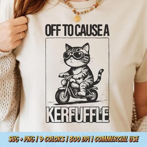 Könnte beinhalten: Cremefarbenes T-Shirt mit einer Schwarz-Weiß-Grafik einer Katze mit Sonnenbrille, die Motorrad fährt. Der Text "OFF TO CAUSE A KERFUFFLE" befindet sich über und unter der Katzenillustration.