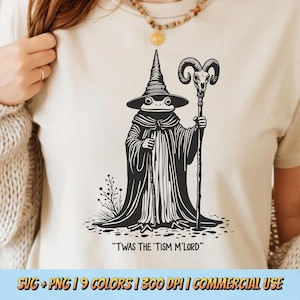 May include: A cream-coloured t-shirt featuring a black and white graphic of a frog wizard holding a staff. The text "TWAS THE 'TISM M'LORD" is below the image. The shirt has a crew neck. The graphic is a whimsical design.